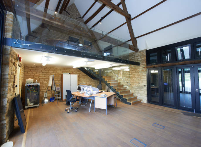 Frameless glass balustrades installed along a mezzanine floor in an open-plan office with exposed beams and stone walls, by Riteweld Engineering.