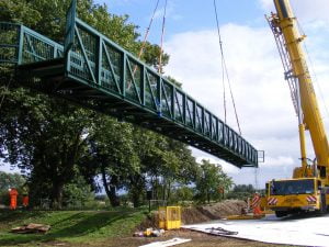 Steel pedestrian bridge being lifted into position by a crane, with construction workers overseeing the installation, as part of a Riteweld Engineering project.