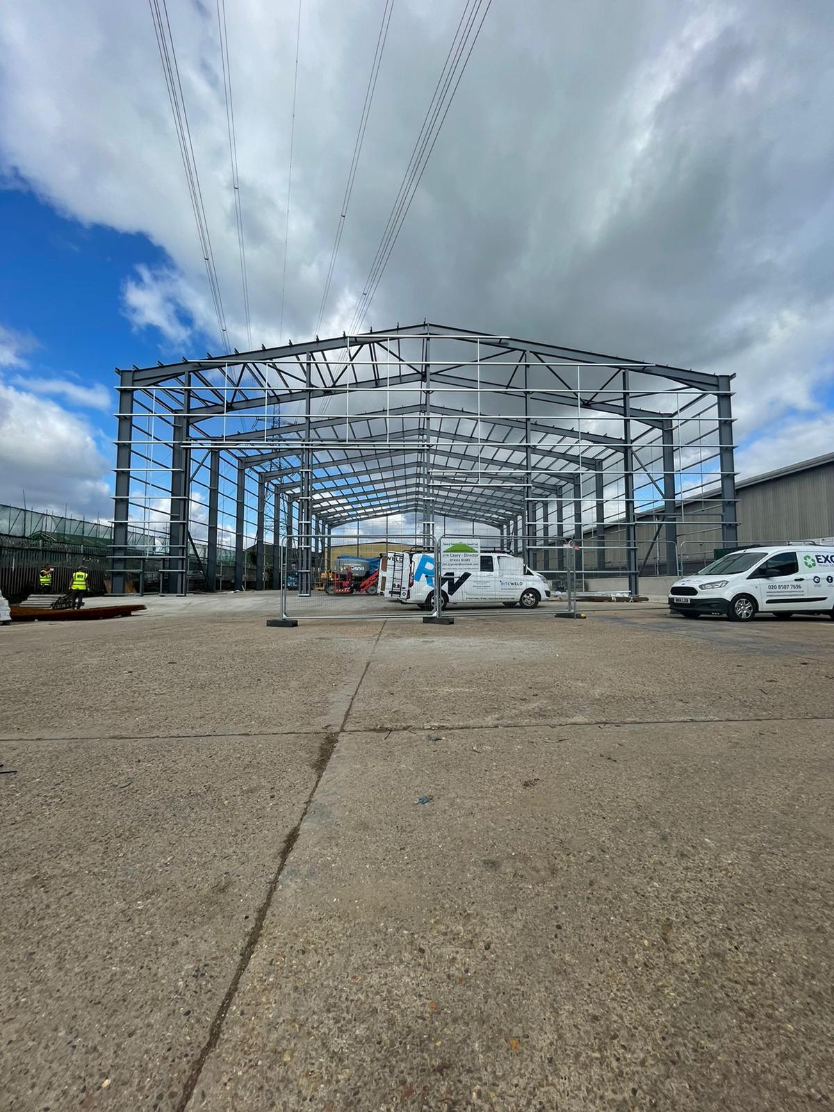 Large structural steel frame being installed for an industrial warehouse, with construction vehicles and Riteweld Engineering vans on site.